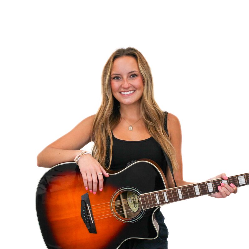 A woman with long hair smiles while holding an acoustic guitar with a sunburst finish. She is wearing a black top and standing against a plain white background.