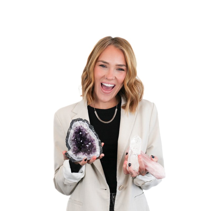 Woman smiling while holding a purple geode in one hand and white crystals in the other, wearing a light-colored blazer.