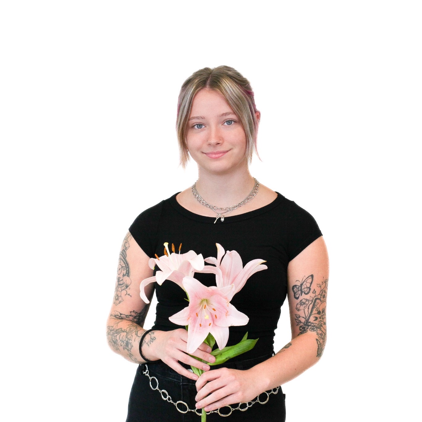 A person with tattoos, wearing a black t-shirt, holding a bouquet of pink lilies, stands against a white background.
