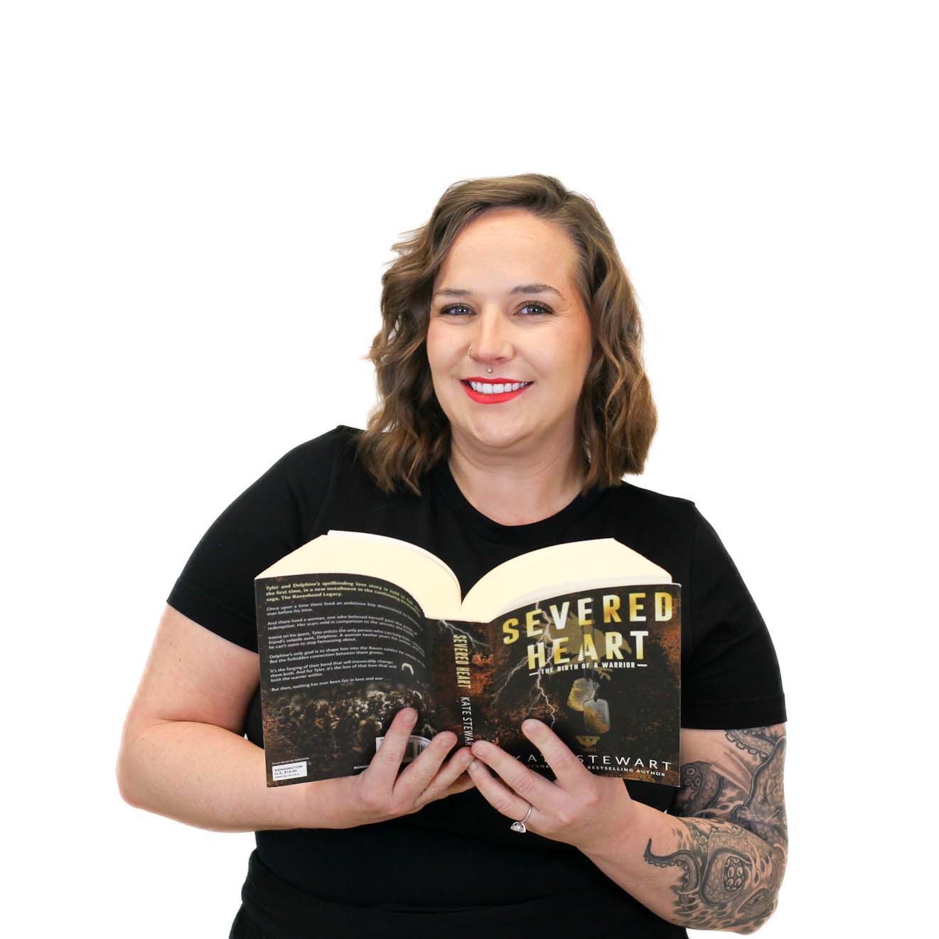 A woman with brown hair and a tattoo on her arm is smiling and holding an open book titled "Severed Heart" against a plain white background.
