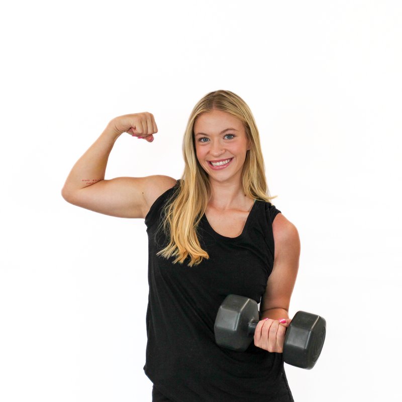 Woman in a black tank top flexes her right arm while holding a dumbbell in her left hand against a white background.