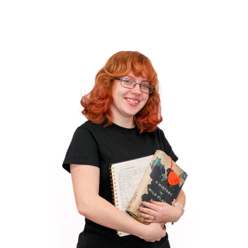 Woman with red hair holding a notebook and a book, smiling at the camera against a white background.