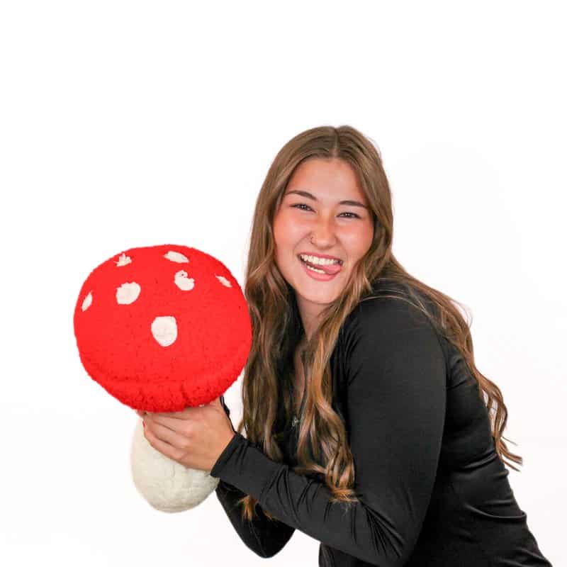 A woman in a black top smiles while holding a large, plush red and white mushroom against a plain white background.