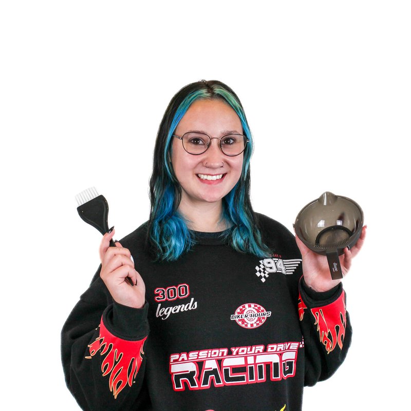 A person with blue hair, wearing glasses and a racing-themed sweater, holds a hair dye brush and bowl, smiling.