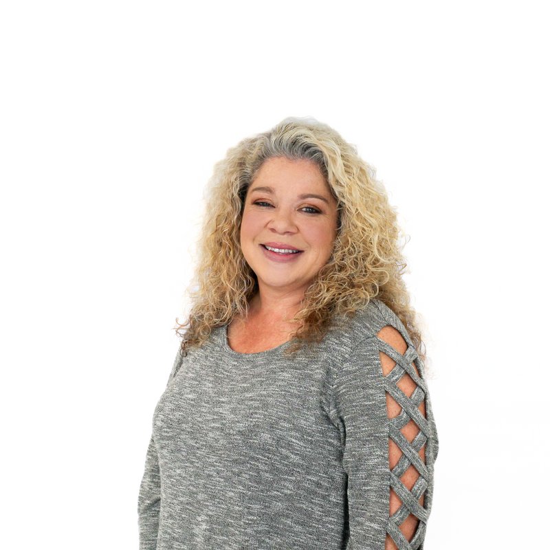 A woman with curly blonde hair wearing a gray long-sleeve top with crisscross sleeve pattern smiles against a white background.