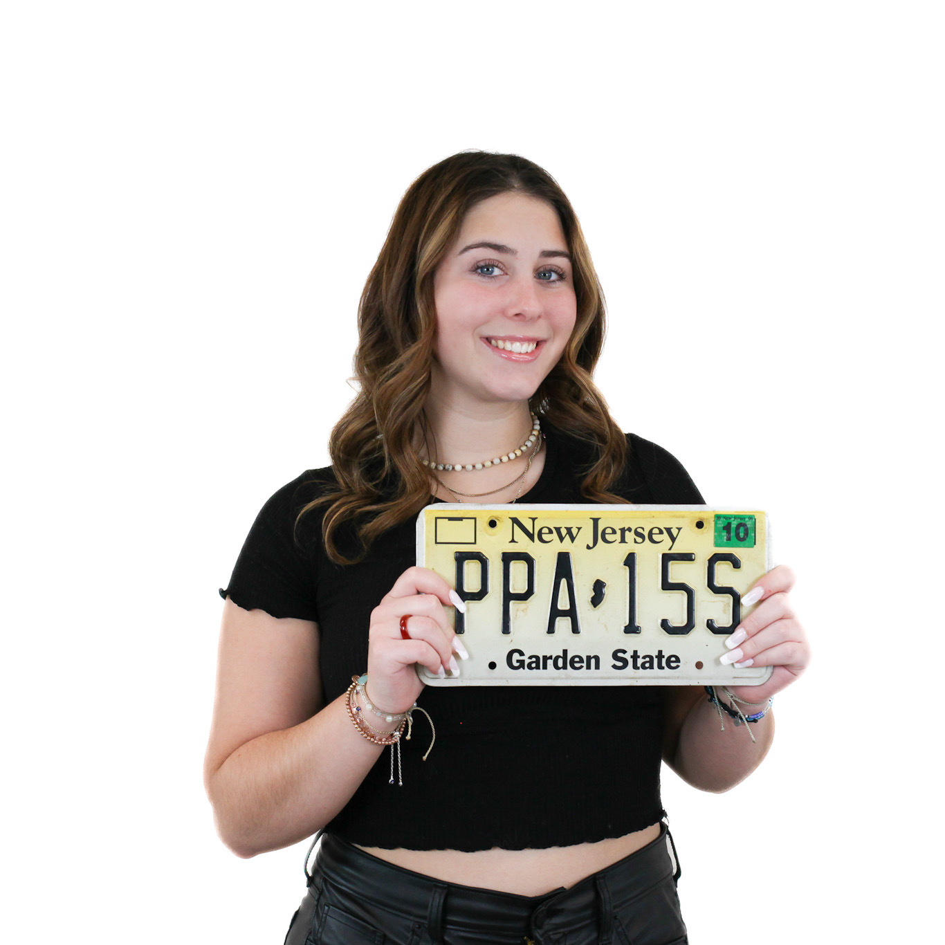 A person with long brown hair, wearing a black shirt, holds a New Jersey license plate with the text "PPA 15S" and smiles at the camera.