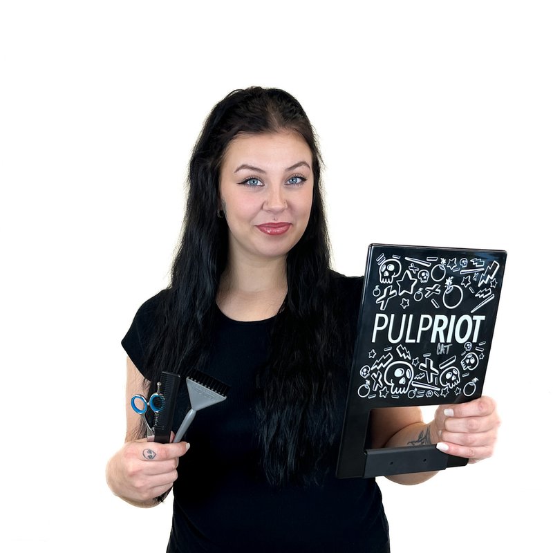 Woman with long dark hair holding hair styling tools and a sign labeled "Pulp Riot," smiling in front of a white background.