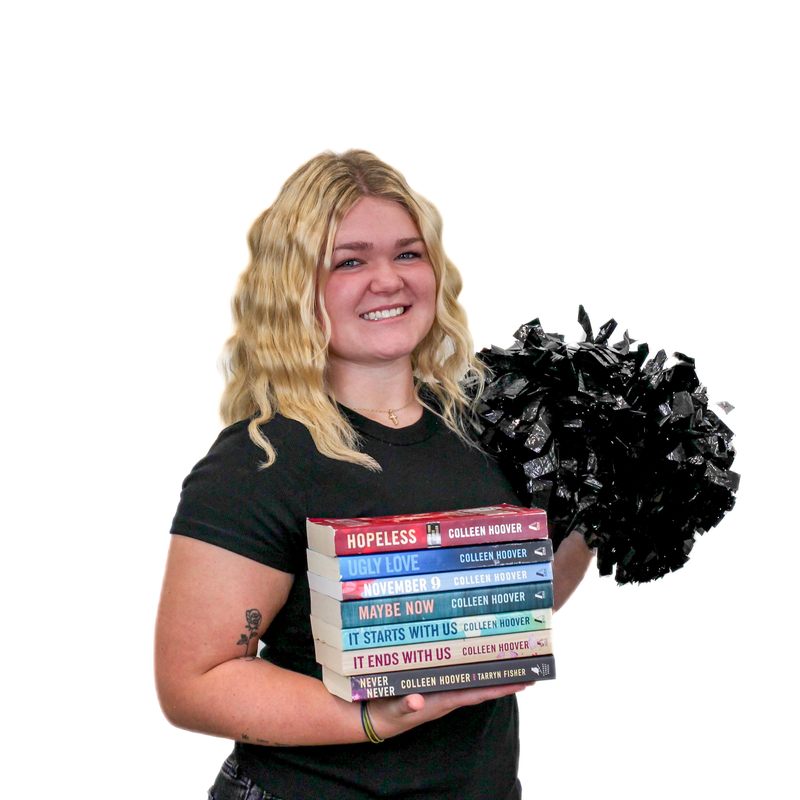 A woman holds a stack of seven books in one hand and a black pom-pom in the other, smiling at the camera.