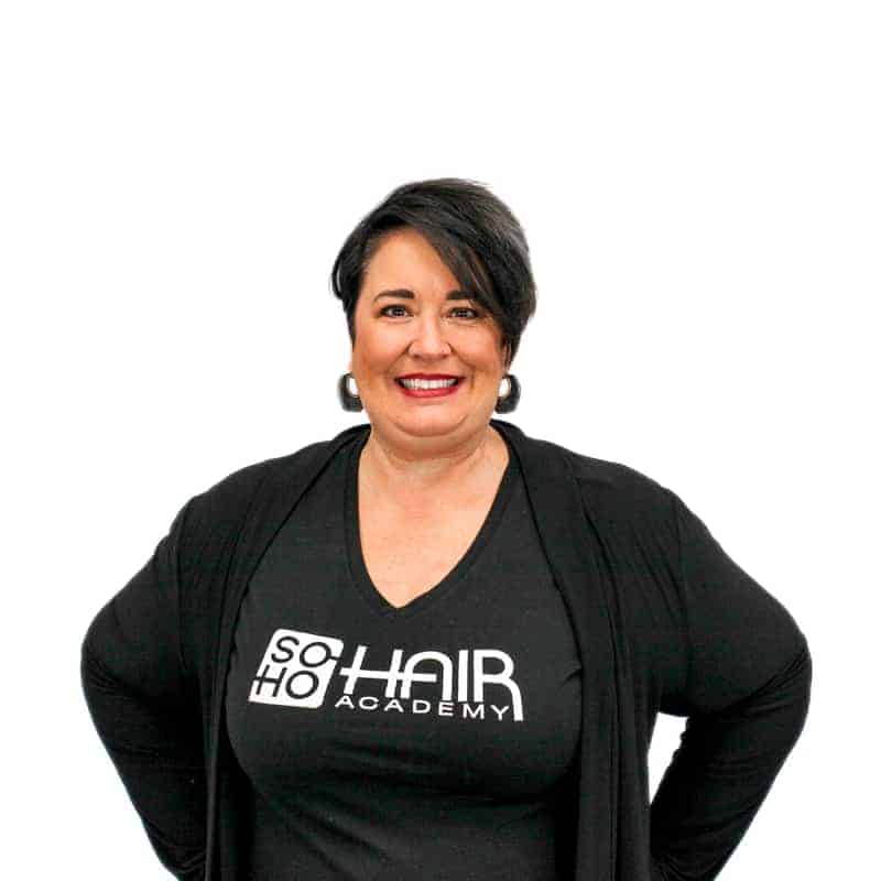 A smiling woman with dark hair dressed in a black outfit from a cosmetology school, featuring its logo on the shirt, standing against a white background.