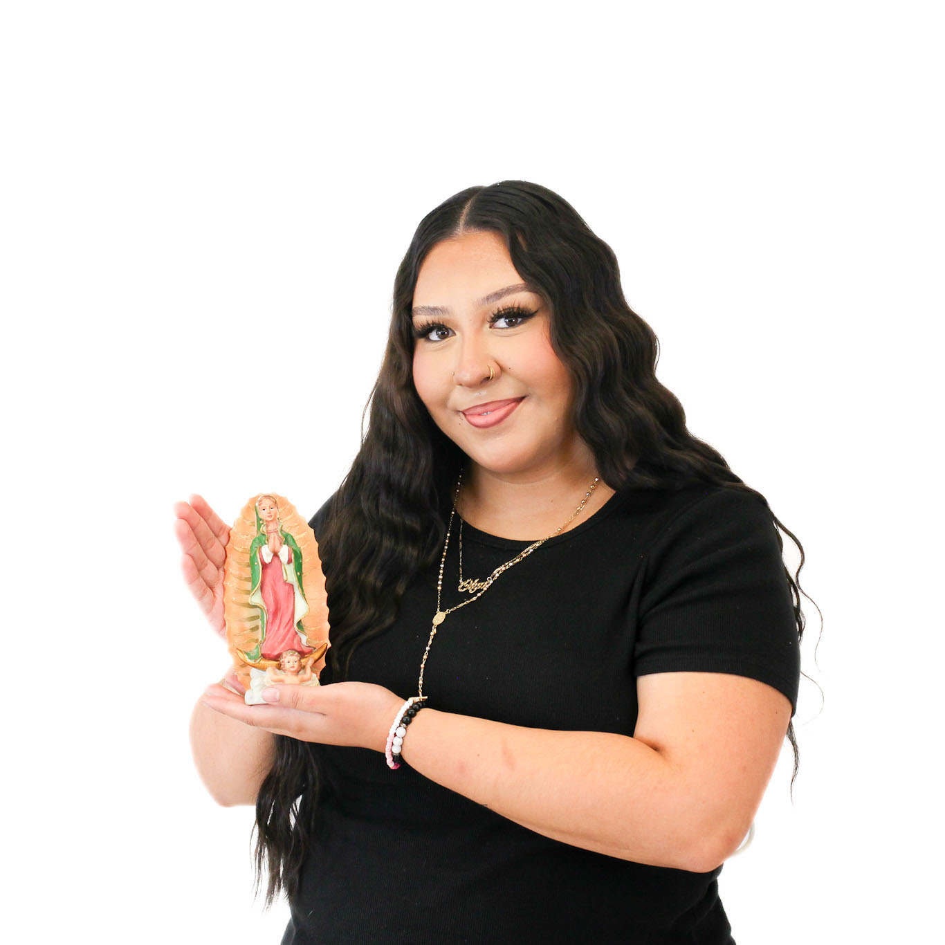 A woman with long dark hair wears a black shirt and holds a small figurine of the Virgin of Guadalupe against a plain white background.