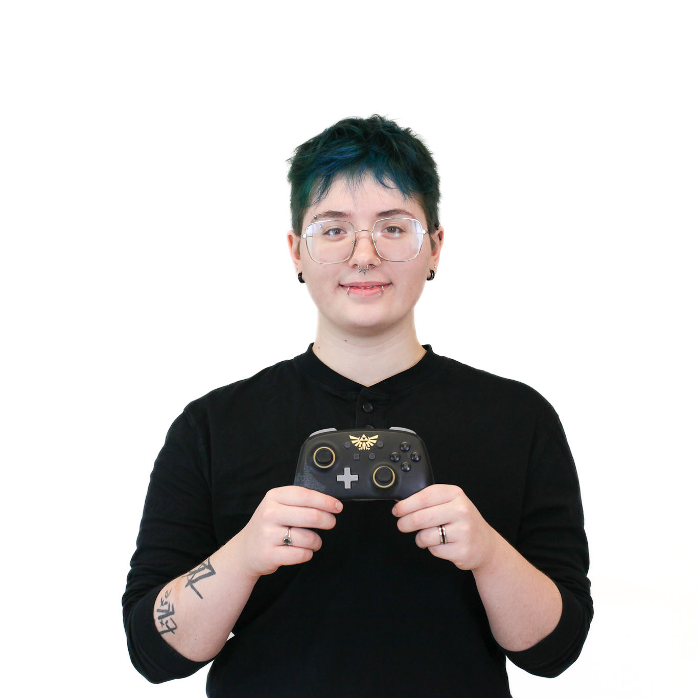 Person with short blue hair and glasses, wearing a black shirt, holding a black game controller with gold accents, standing against a white background.