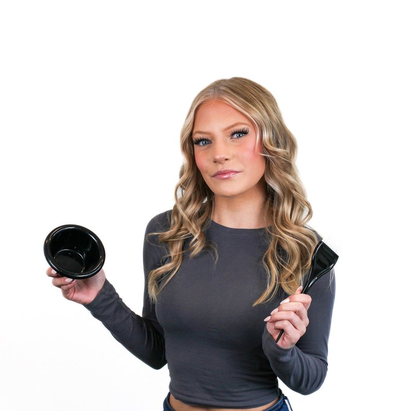 Woman with long, wavy hair holds a black bowl in one hand and a brush in the other, against a white background.