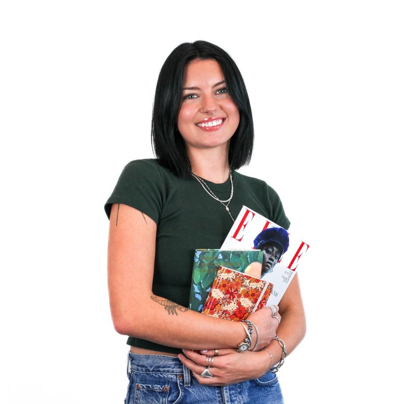 A person with dark hair, wearing a green t-shirt and jeans, smiling while holding several magazines, including one with the title "Elle.