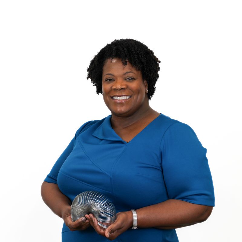 A person wearing a blue dress stands holding a metal slinky. They are smiling and positioned against a plain white background.