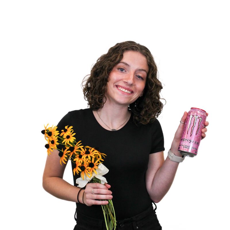 A person with curly hair is smiling while holding a bouquet of yellow flowers in one hand and a pink can of Monster energy drink in the other hand.