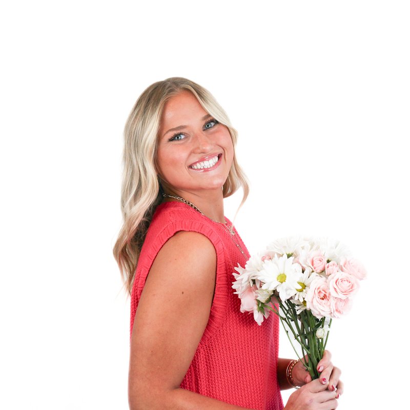 Smiling woman in a pink sleeveless top holding a bouquet of pink and white flowers against a white background.