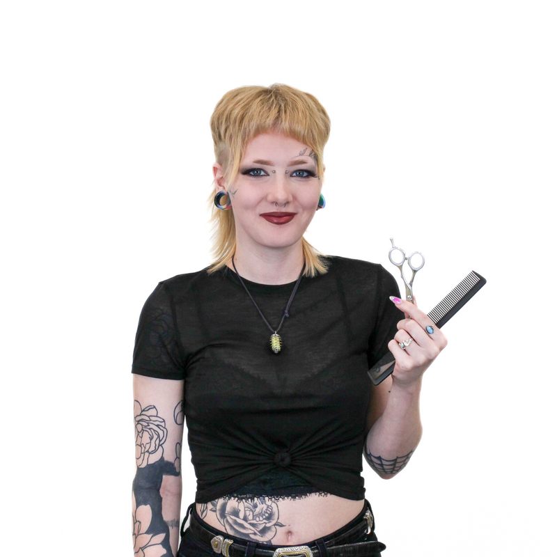 A person with facial piercings and tattoos holds a pair of scissors and a comb, wearing a black crop top and pants.