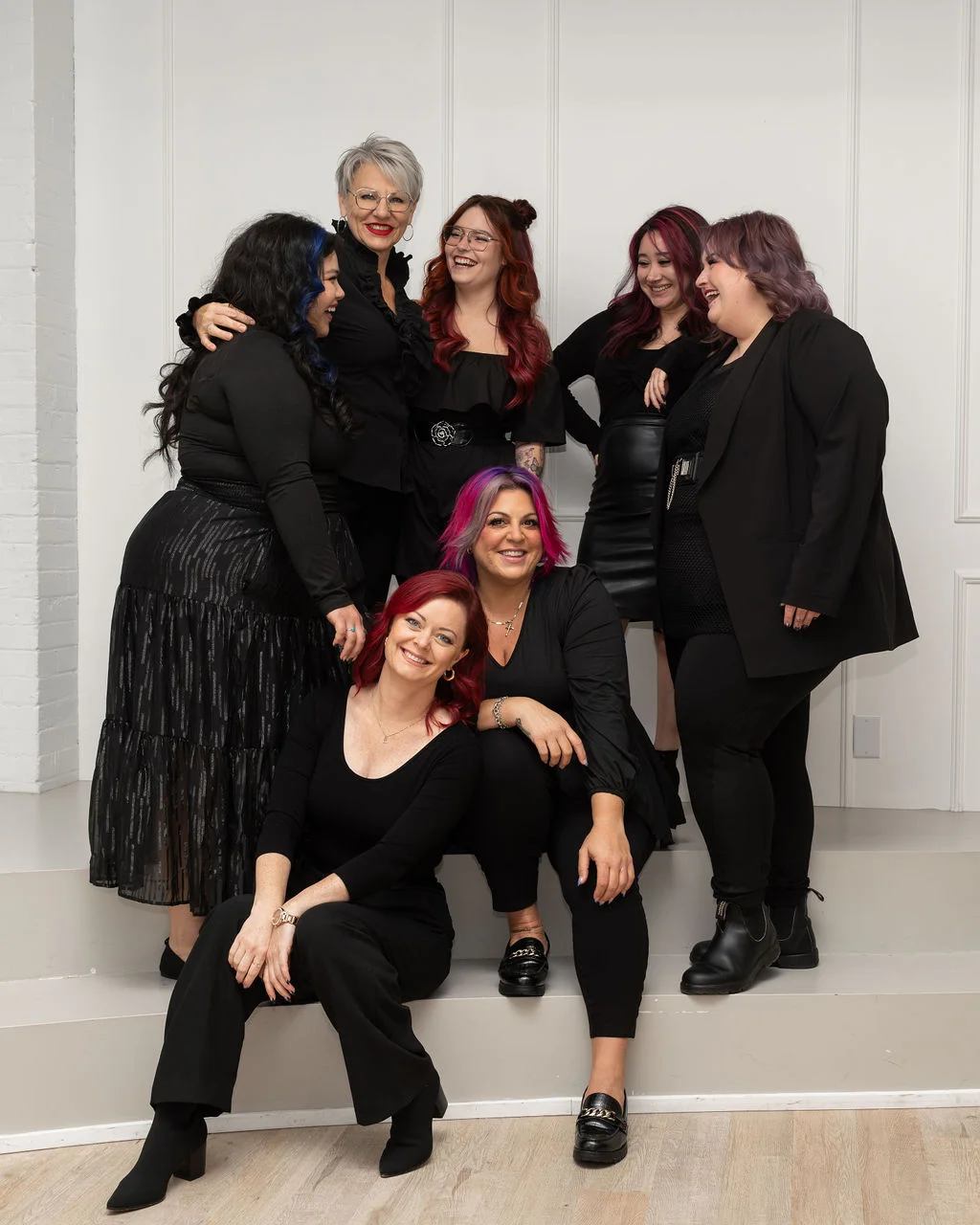 A group of seven smiling women in black attire, fresh from a cosmetology school, posing together indoors.