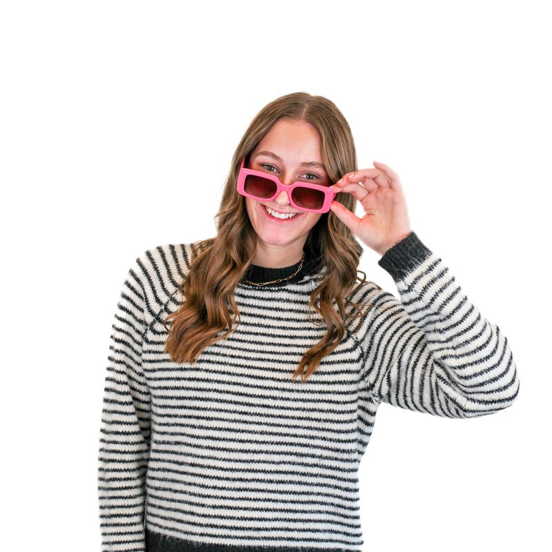 Woman with long hair wearing a striped sweater and pink sunglasses, smiling while holding the sunglasses with one hand against a white background.