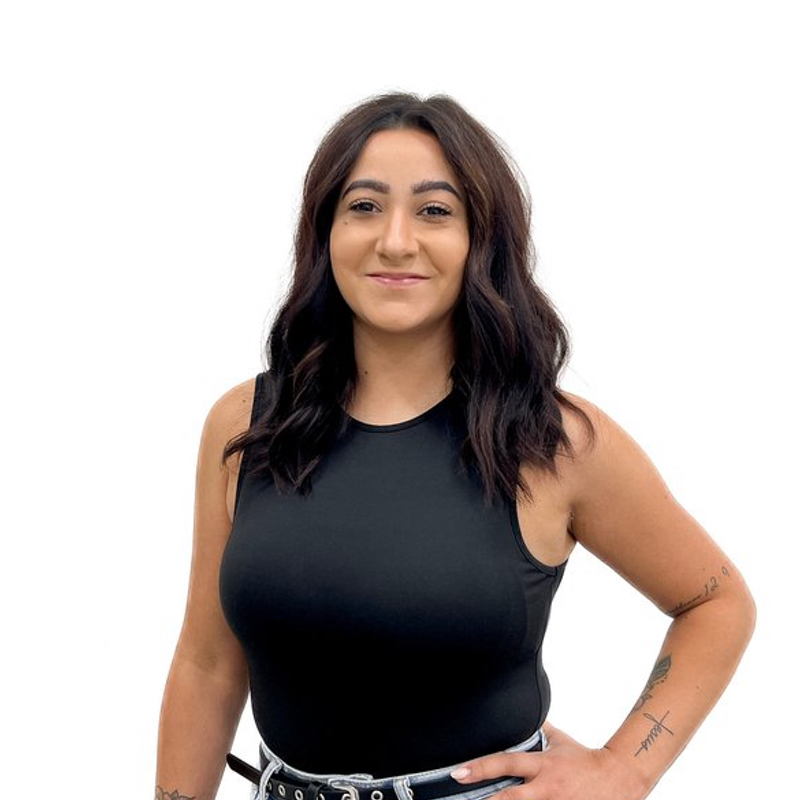 A woman with wavy dark hair wearing a black sleeveless top and black pants stands against a white background. She has tattoos on her arms and a slight smile on her face.