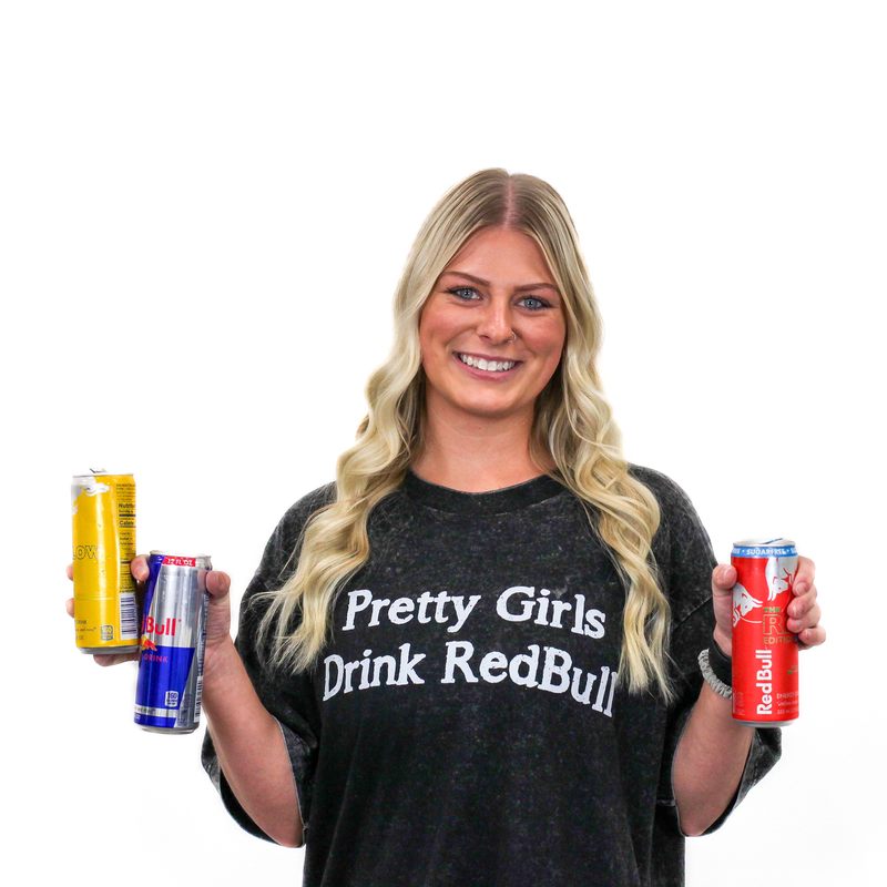 A person with long blonde hair smiles and holds three cans of Red Bull energy drink, wearing a black shirt with the text "Pretty Girls Drink Red Bull.