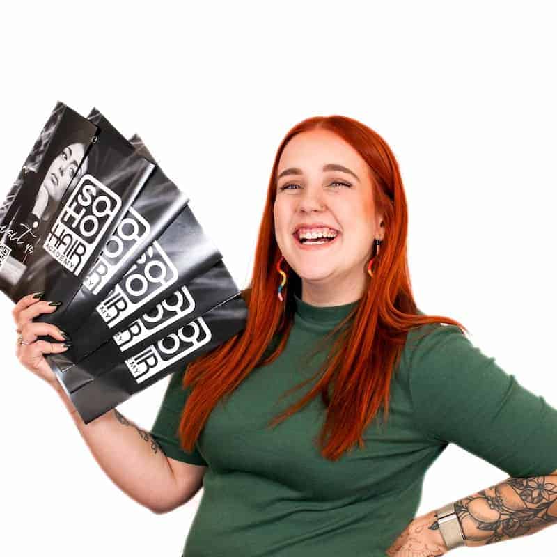 A cheerful woman with red hair, recently graduated from cosmetology school, holding shopping bags and smiling at the camera.