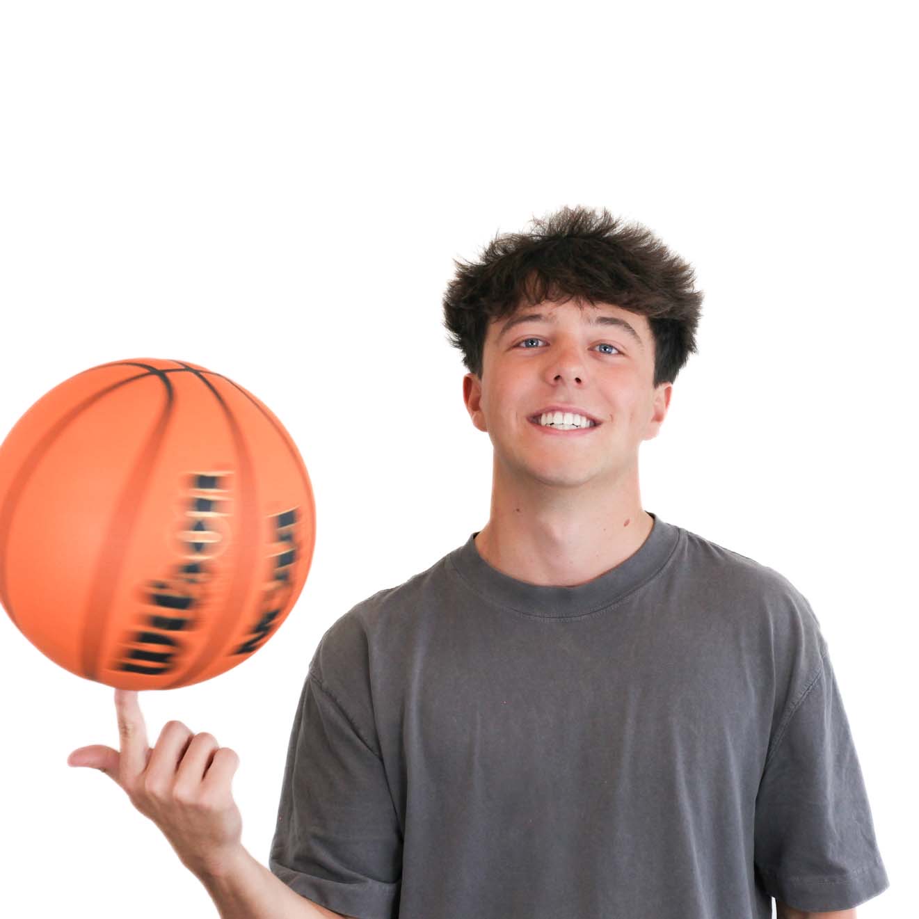A young man in a gray t-shirt smiles while spinning an orange basketball on his finger against a plain white background.