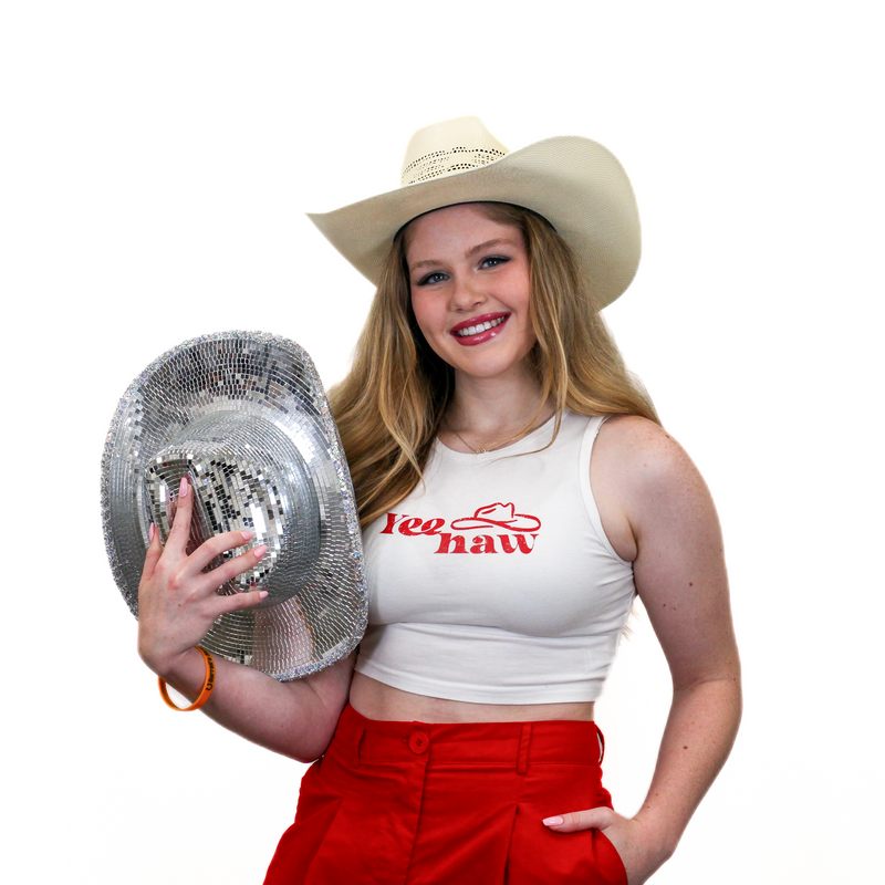 A woman in a white cowboy hat and a "Yee Haw" tank top smiles while holding a silver sequin cowboy hat. She is wearing red pants and standing against a white background.