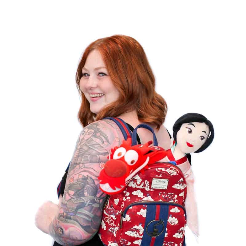Woman with tattooed arm and beautiful hair smiling over her shoulder, carrying a backpack with plush toys attached.