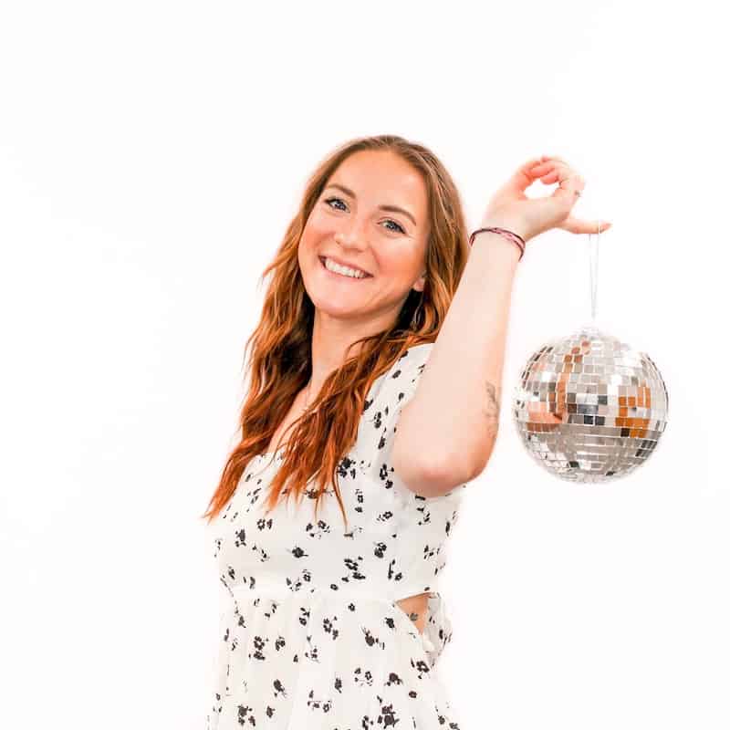 A smiling woman holding a disco ball with one hand against a white background at a beauty salon.