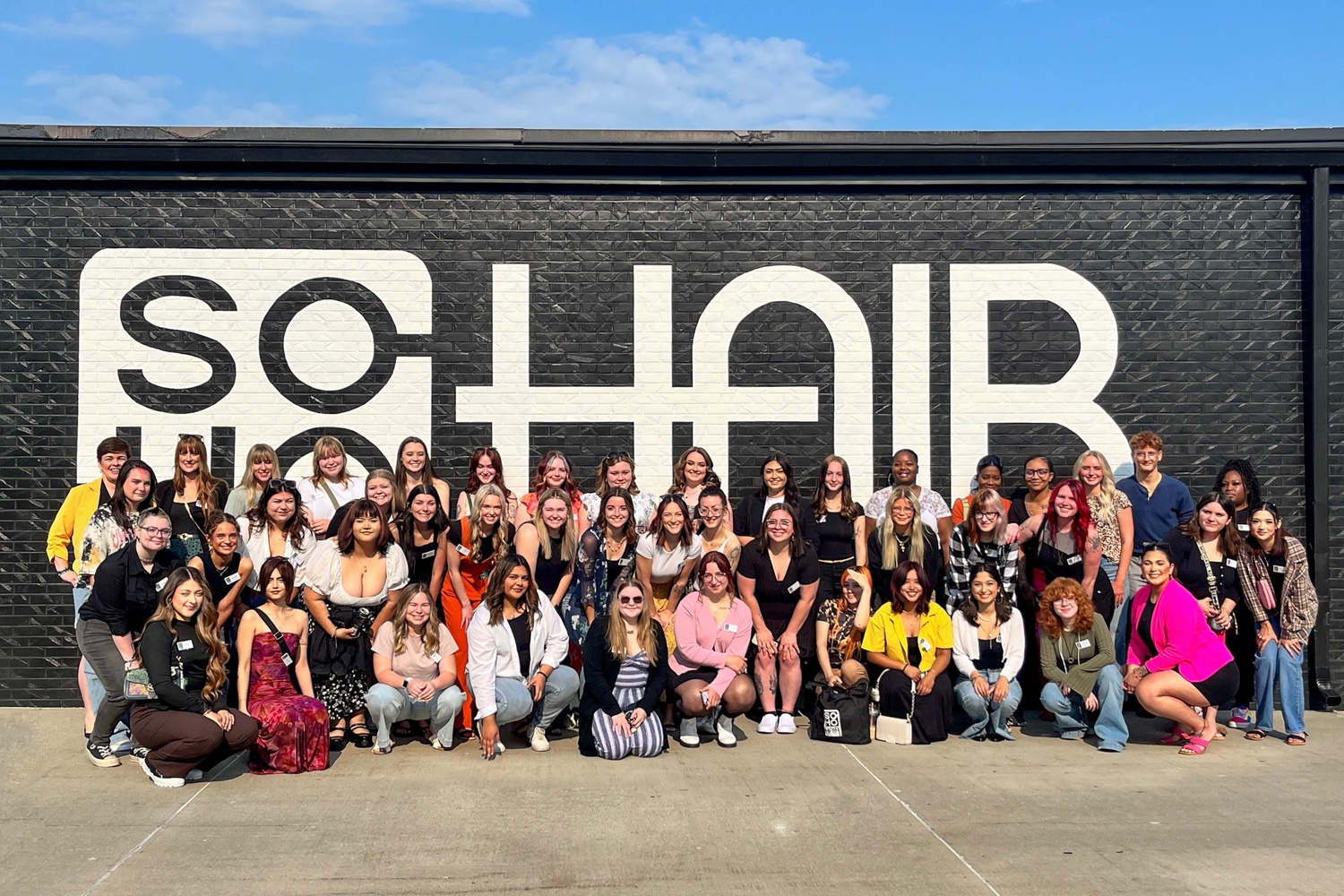 A group of individuals pose together in front of a large mural with the word "SOHAIR" painted on a brick wall.