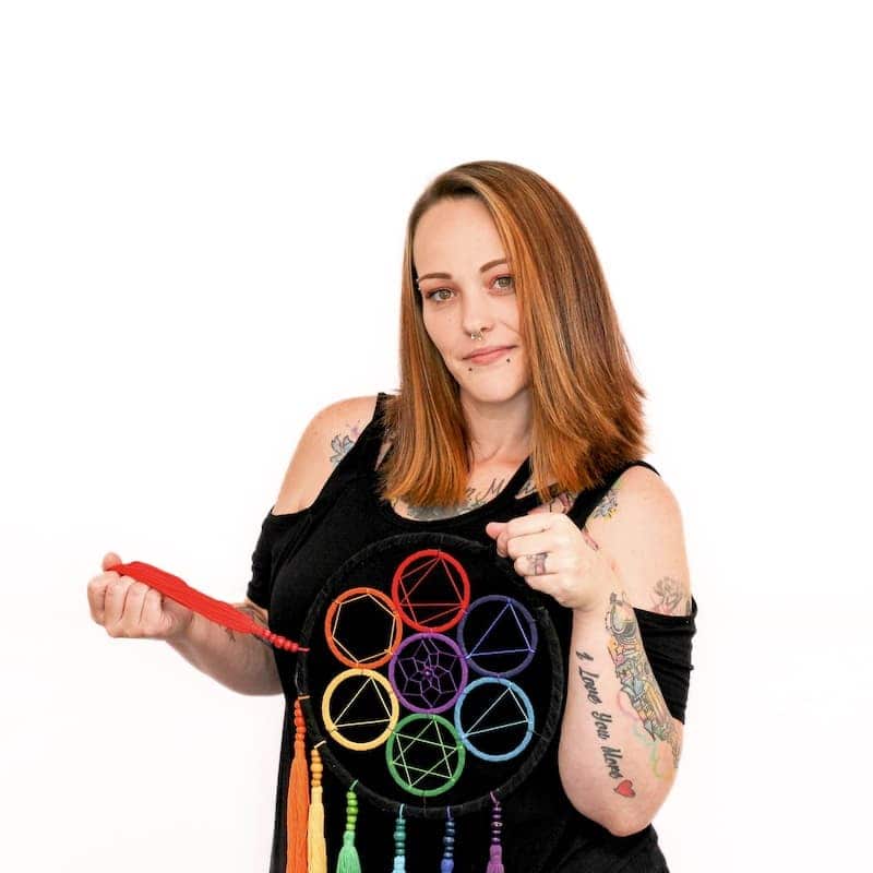 A woman with tattoos, fresh from the hair salon, holds a colorful dreamcatcher and looks at the camera with a slight smile.