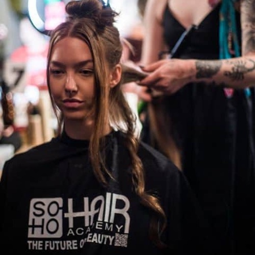 A person gets their hair styled at a salon. They are wearing a black cape with "Soho Hair Academy" printed on it.